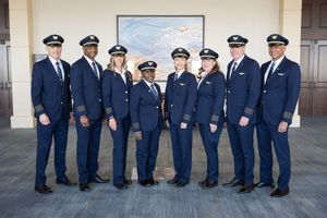 Group of eight airline pilots in navy uniforms posing together indoors for a professional photo.