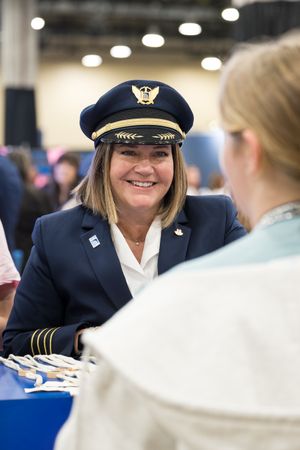 Female airline pilot in uniform and captain's hat smiles at a woman during an aviation career fair event.
