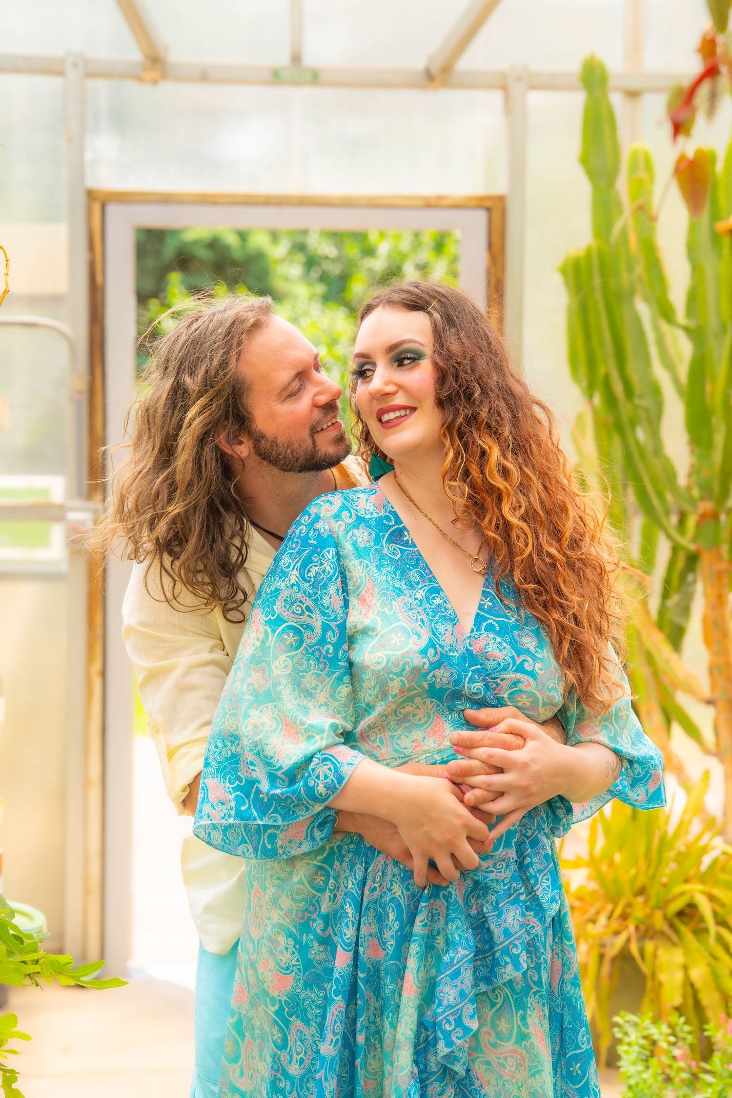 A couple embraces in a greenhouse setting with tropical plants, the woman wearing a flowing turquoise patterned dress.