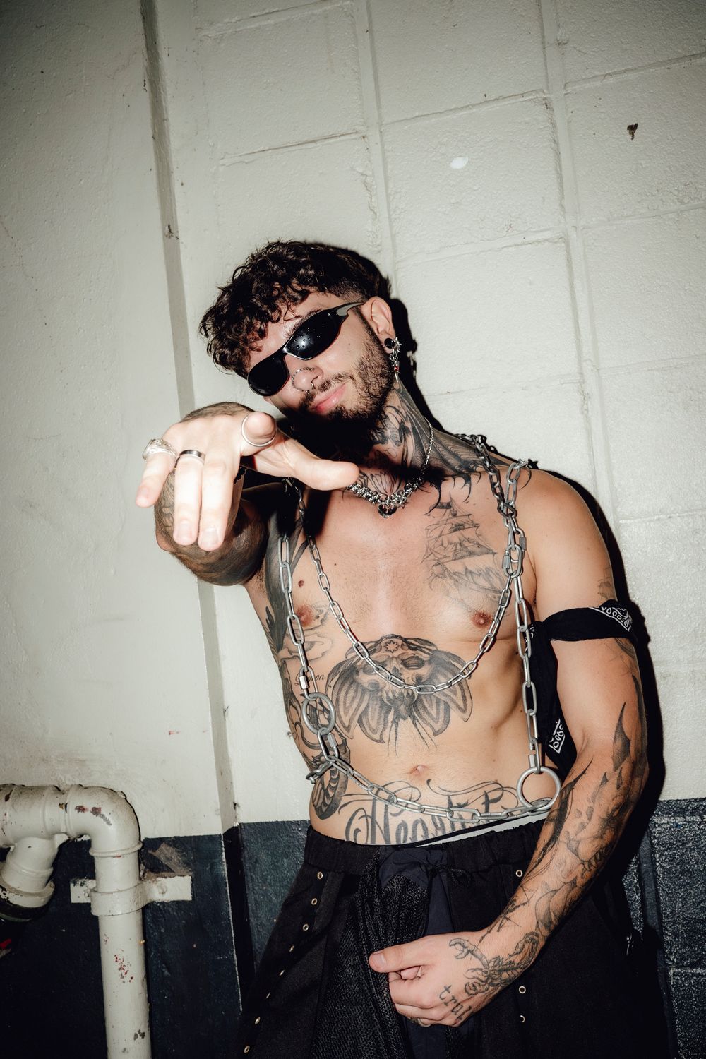 Tattooed person in sunglasses gesturing against white wall with industrial pipe.
