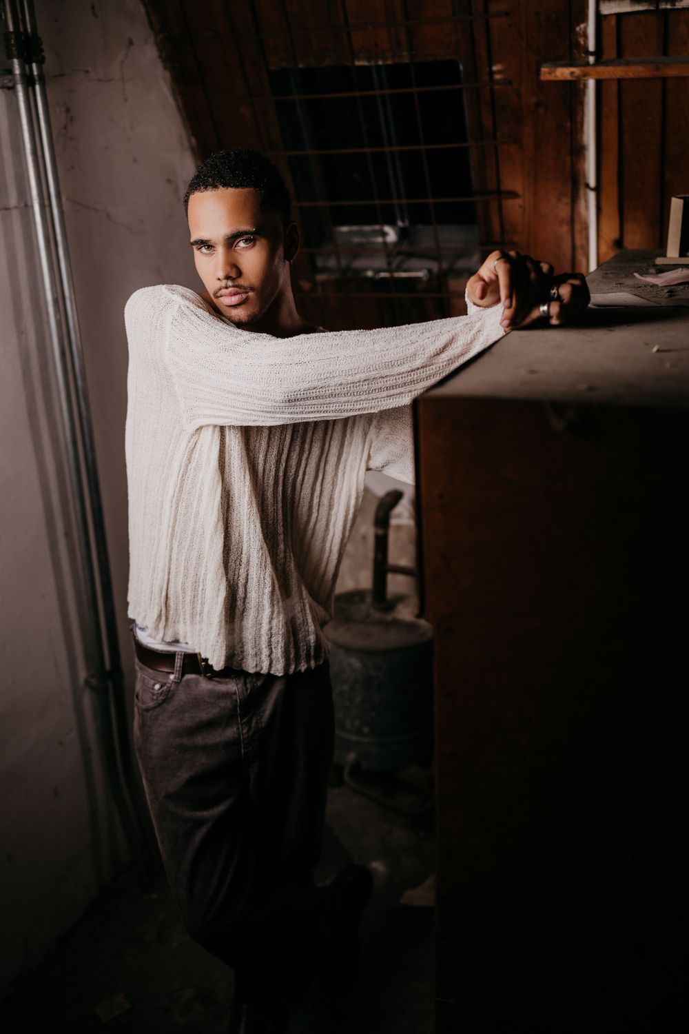Person in cream sweater and dark pants poses dramatically against rustic industrial backdrop in moody lighting.