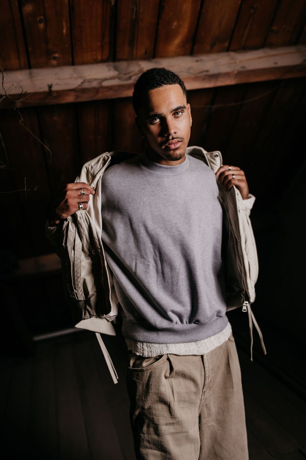 A person in a light gray sweatshirt and beige pants poses in a dimly lit setting.