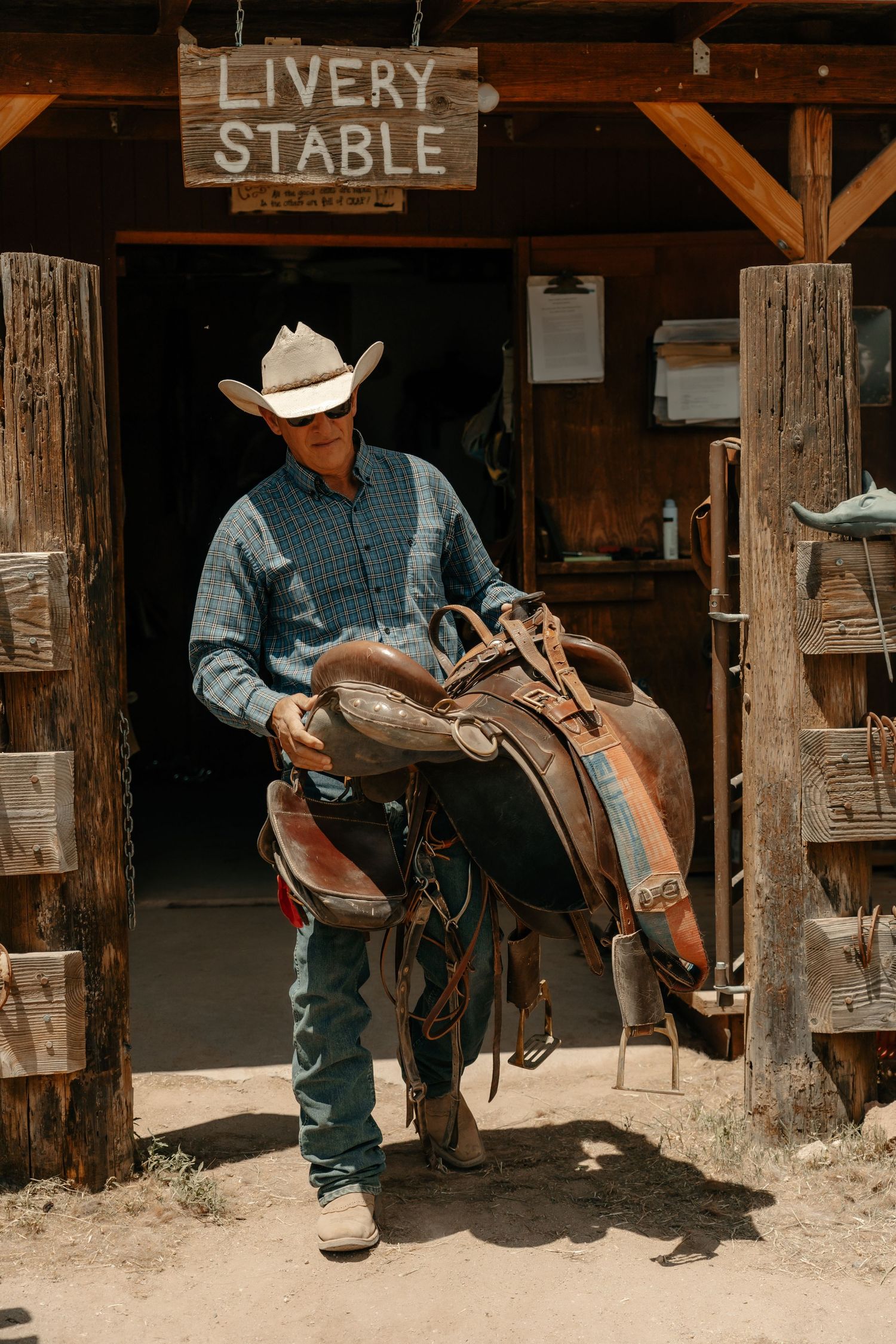 A rancher inspects horse tack under a wooden Livery Stable sign.