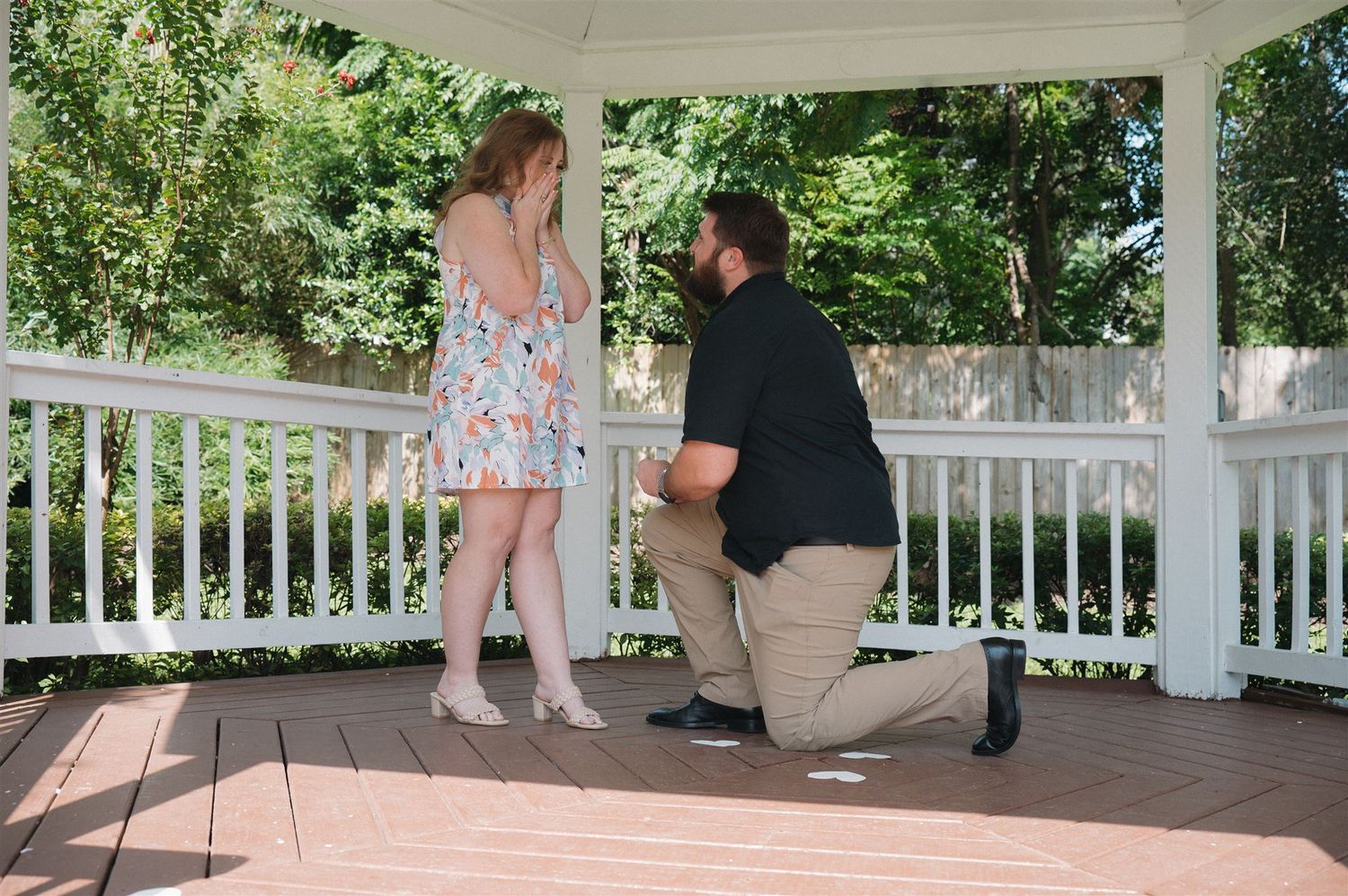 A person kneels down to propose on a white porch surrounded by green foliage on a summer day.