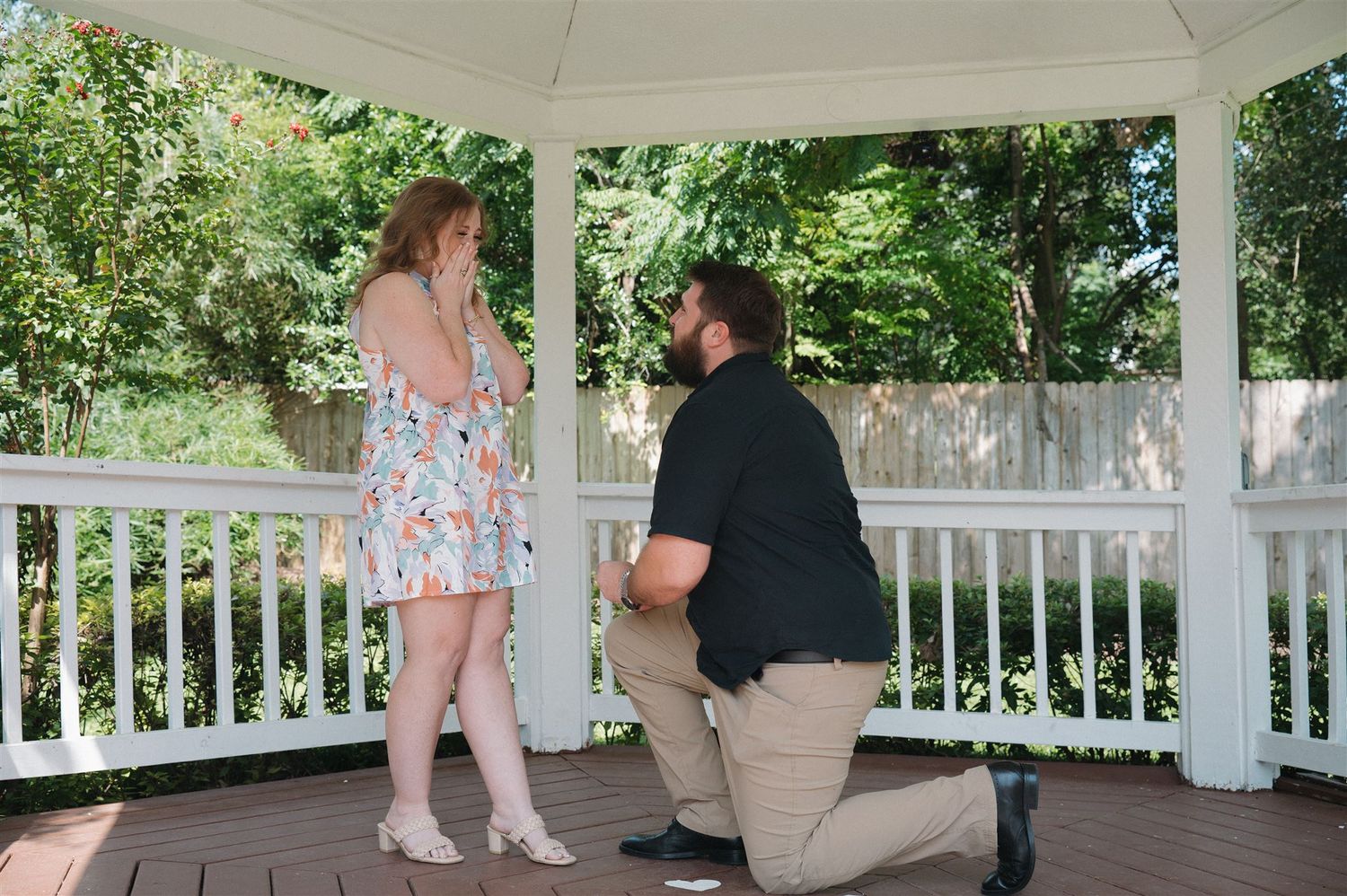 A romantic proposal moment in a white wooden gazebo surrounded by lush summer greenery and trees.