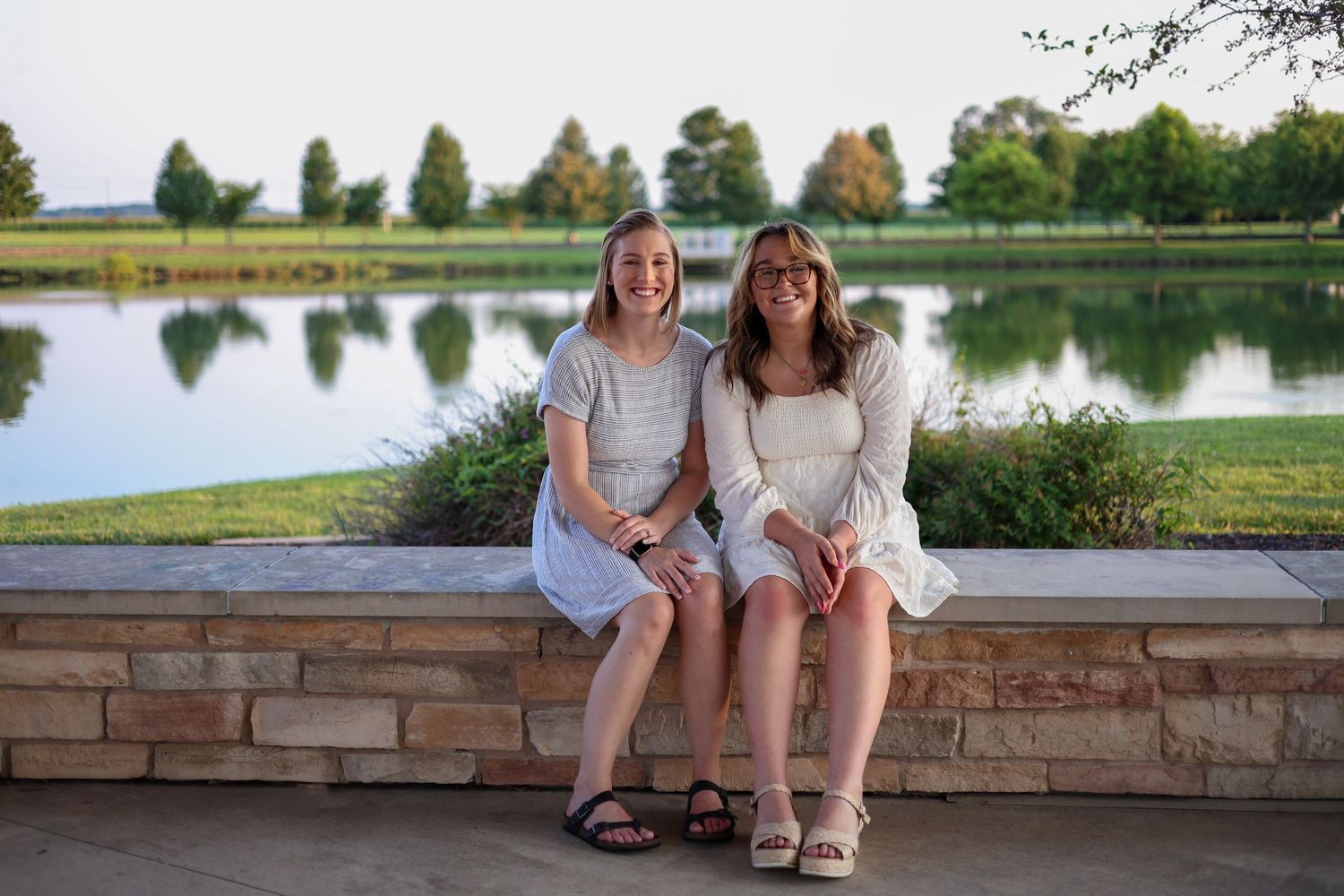 Two people sit together on stone wall near serene lake at sunset.