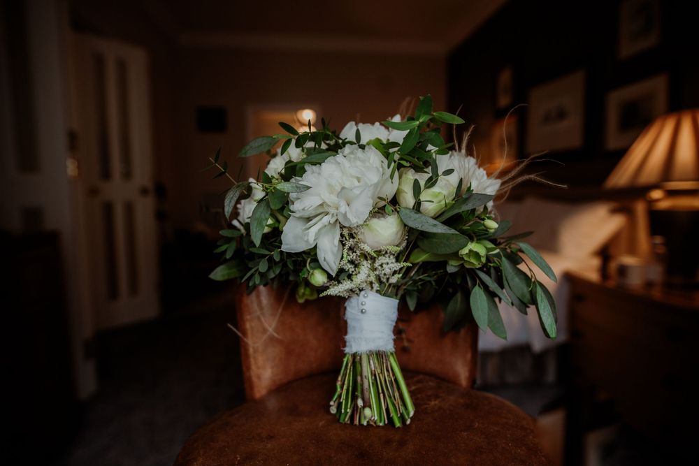 White floral wedding bouquet with greenery sits on brown leather chair.