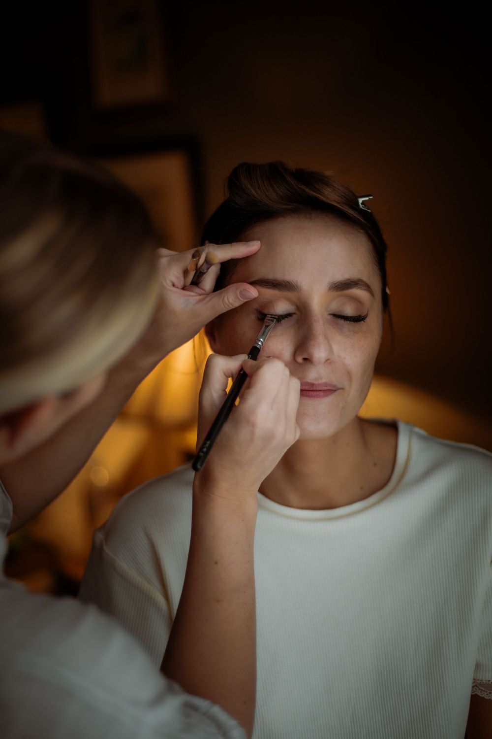 A makeup artist carefully applying eye makeup in warm, moody lighting.