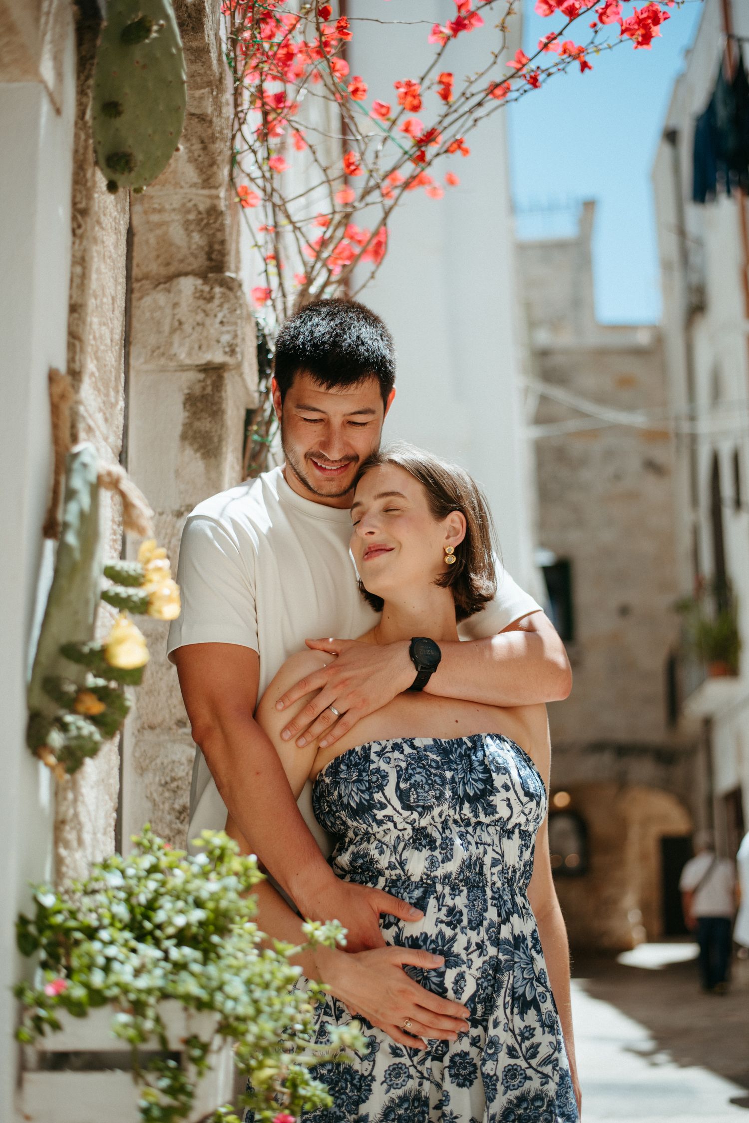 A couple embraces joyfully beneath blooming red flowers against white stone walls.