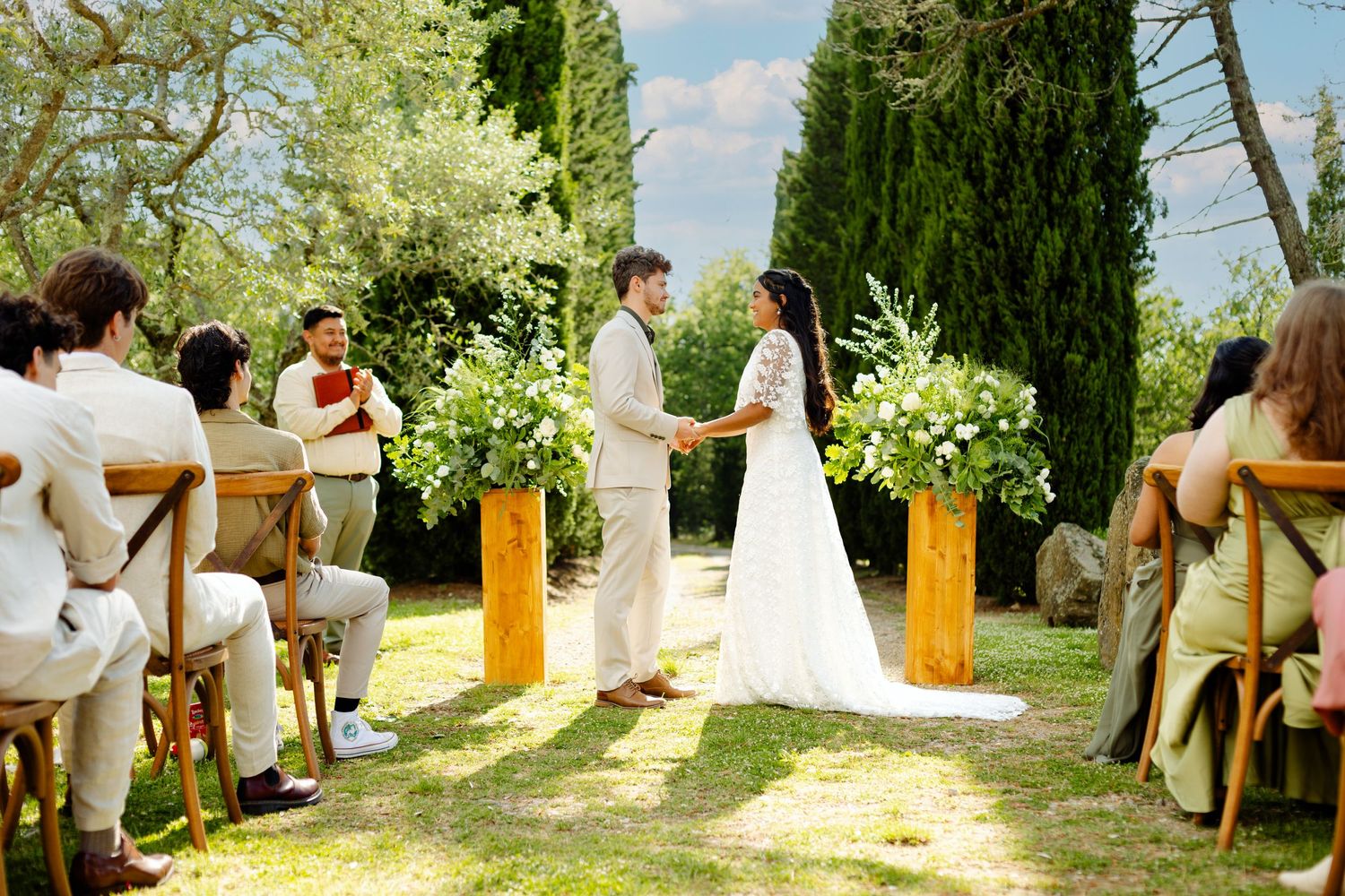 An outdoor wedding ceremony with guests seated on wooden chairs amid lush greenery and cypress trees.