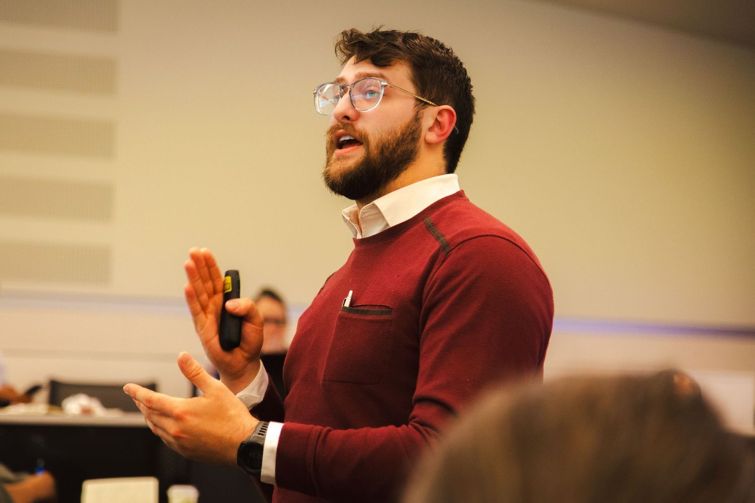 A person in a burgundy sweater gestures animatedly while speaking to an audience in an indoor setting.