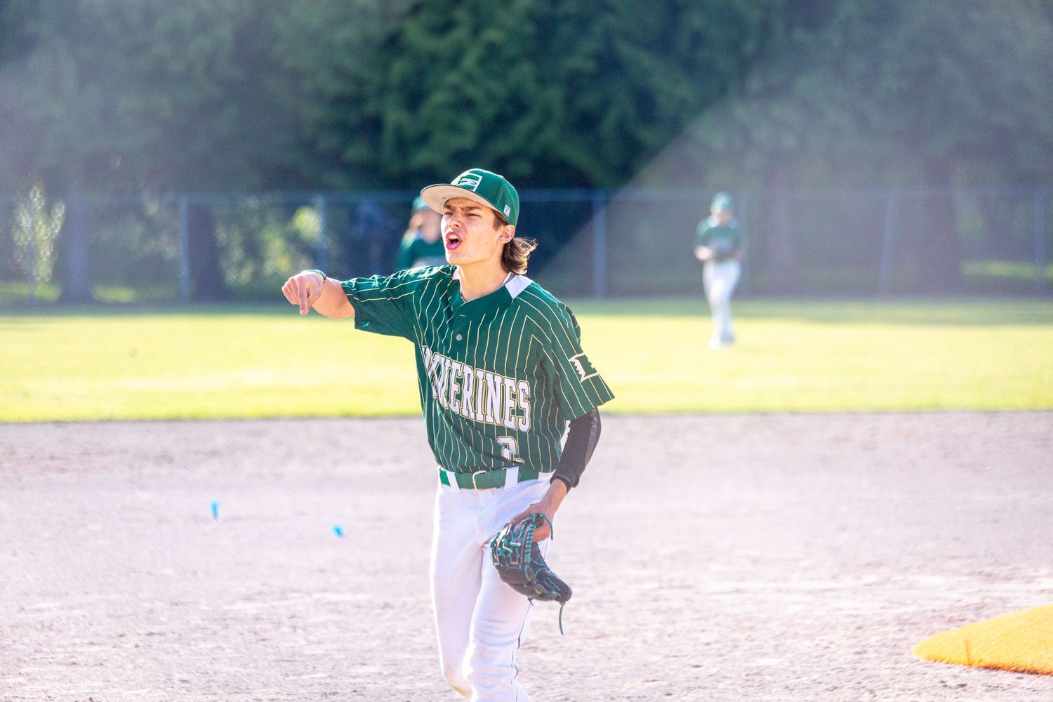 A youth baseball player in a green Keltronics uniform winds up to pitch on a sunny day at the baseball field.