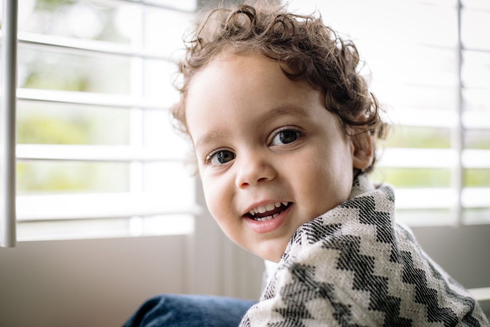 Portrait of a smiling child with curly hair wearing a chevron patterned sweater near a window.