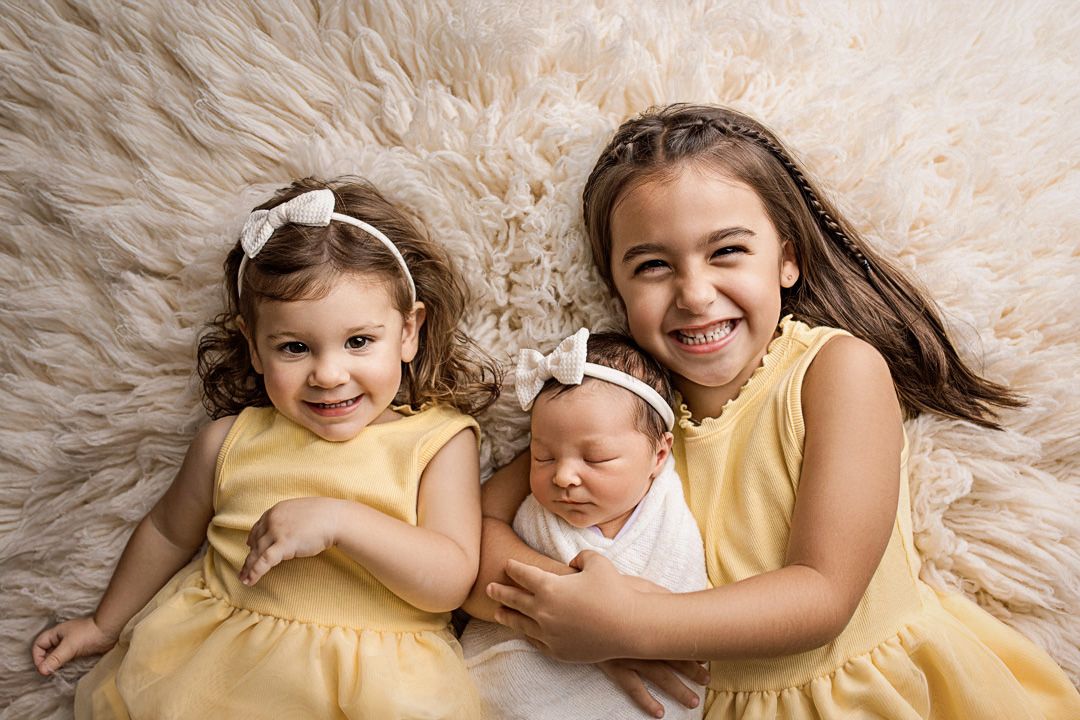 Three siblings in yellow and white dresses laying together on a cream-colored flokati during a newborn photo session in cedar park tx