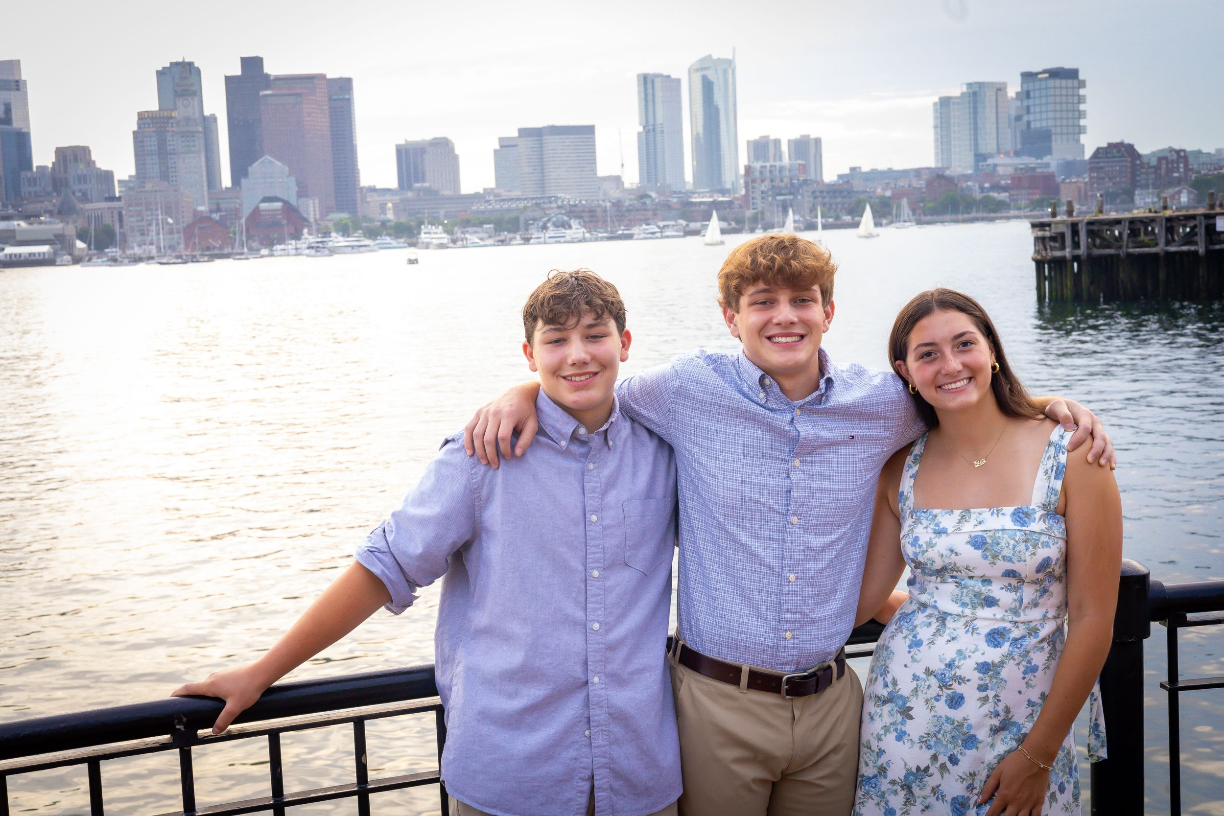 Three people smile together on a waterfront with Boston harbor and skyline in the background.
