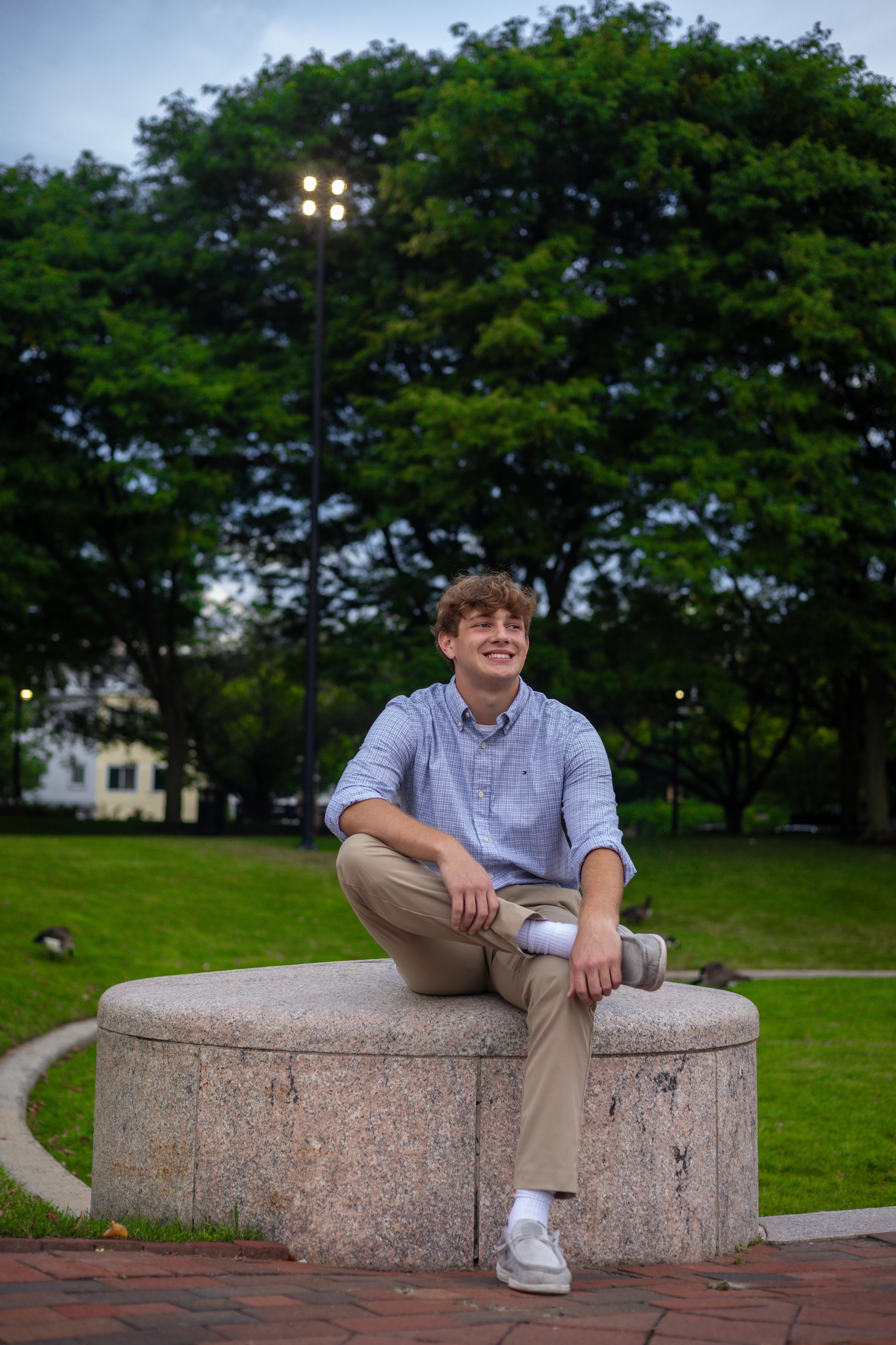 A person in casual attire sits on a stone bench in a park at dusk with trees in the background.
