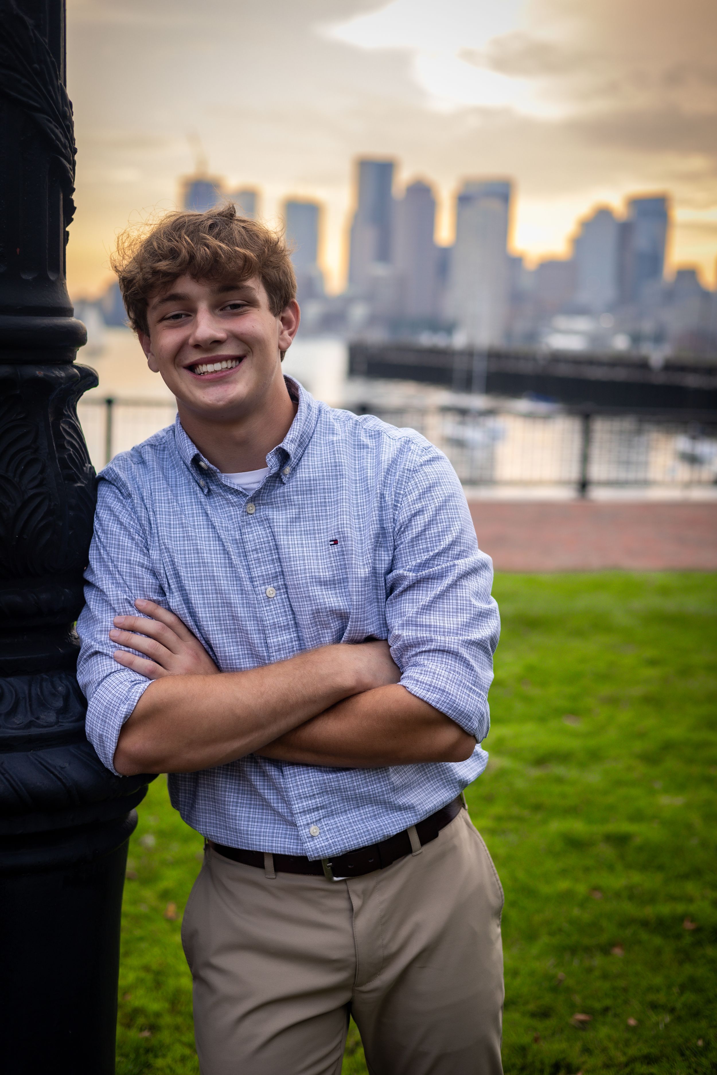 A person poses against a lamp post with Boston skyline at sunset in the background.