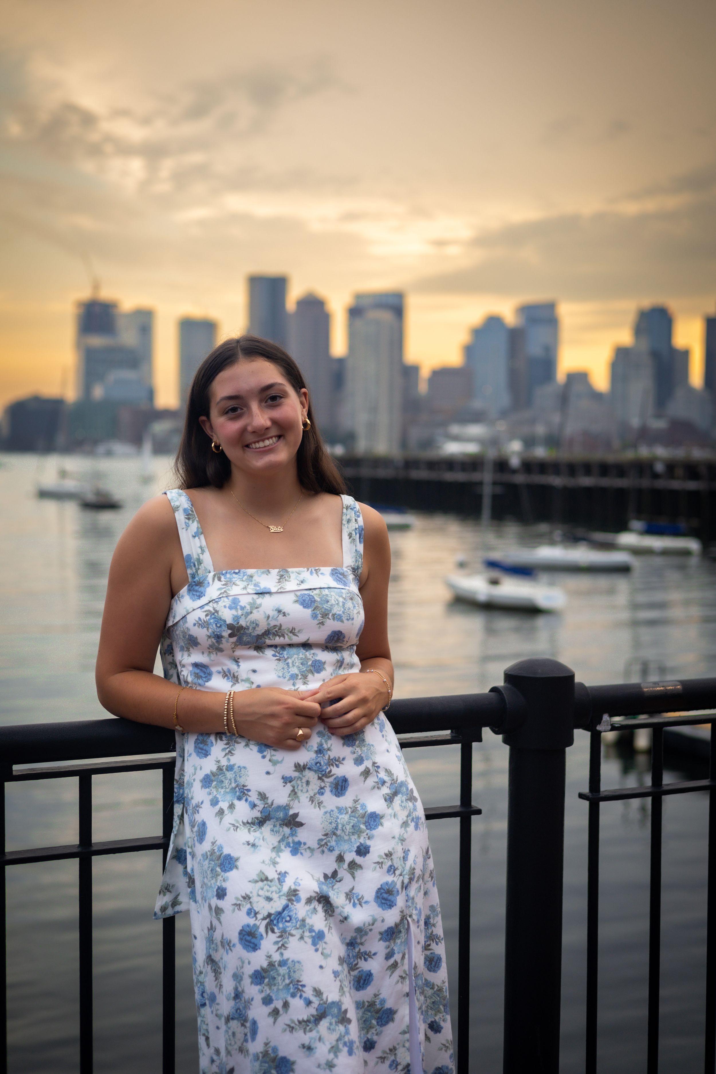 A woman in a floral dress stands by a harbor railing with city skyline and boats in the background.