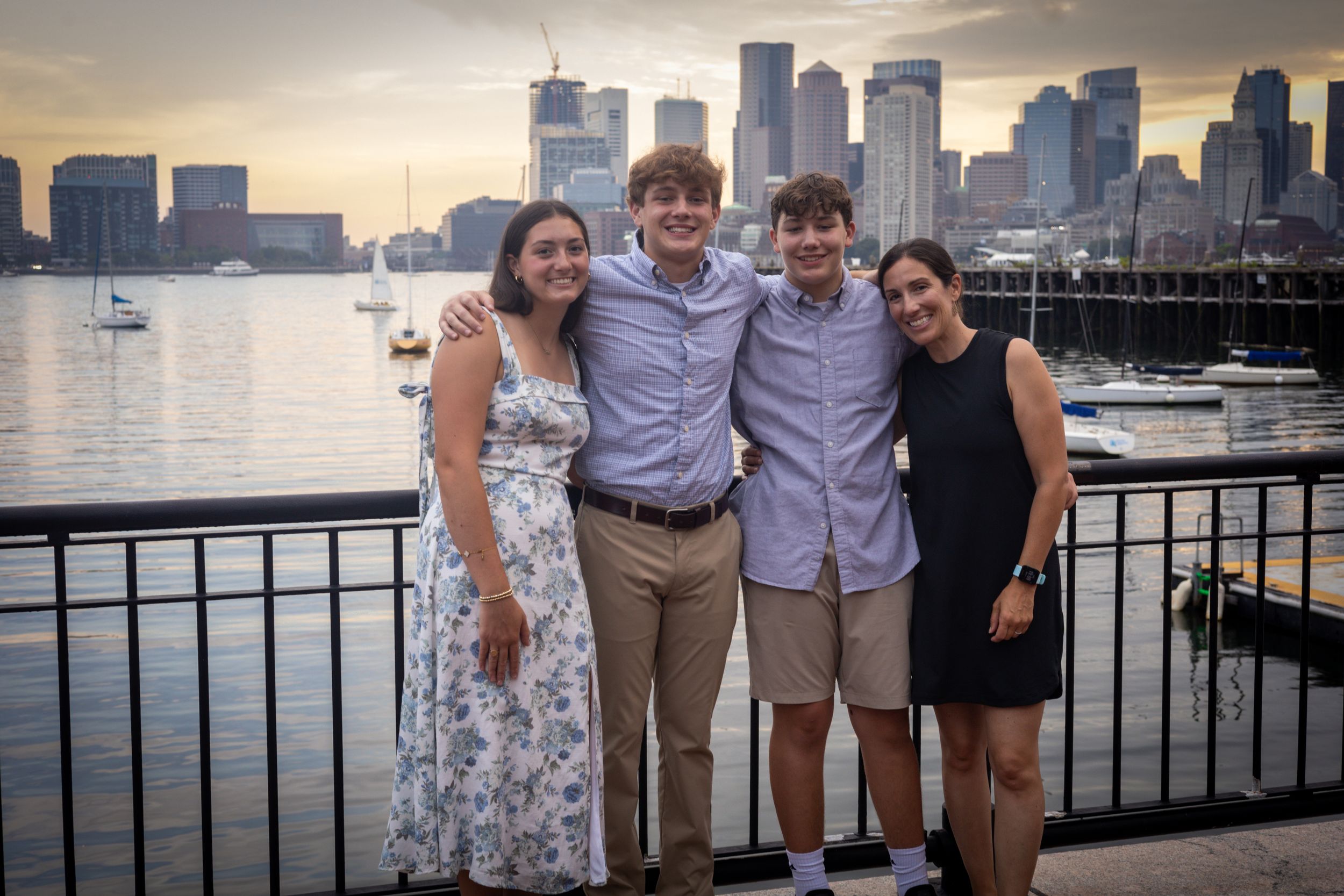 A group poses together on a waterfront boardwalk with Boston harbor and skyline at sunset.