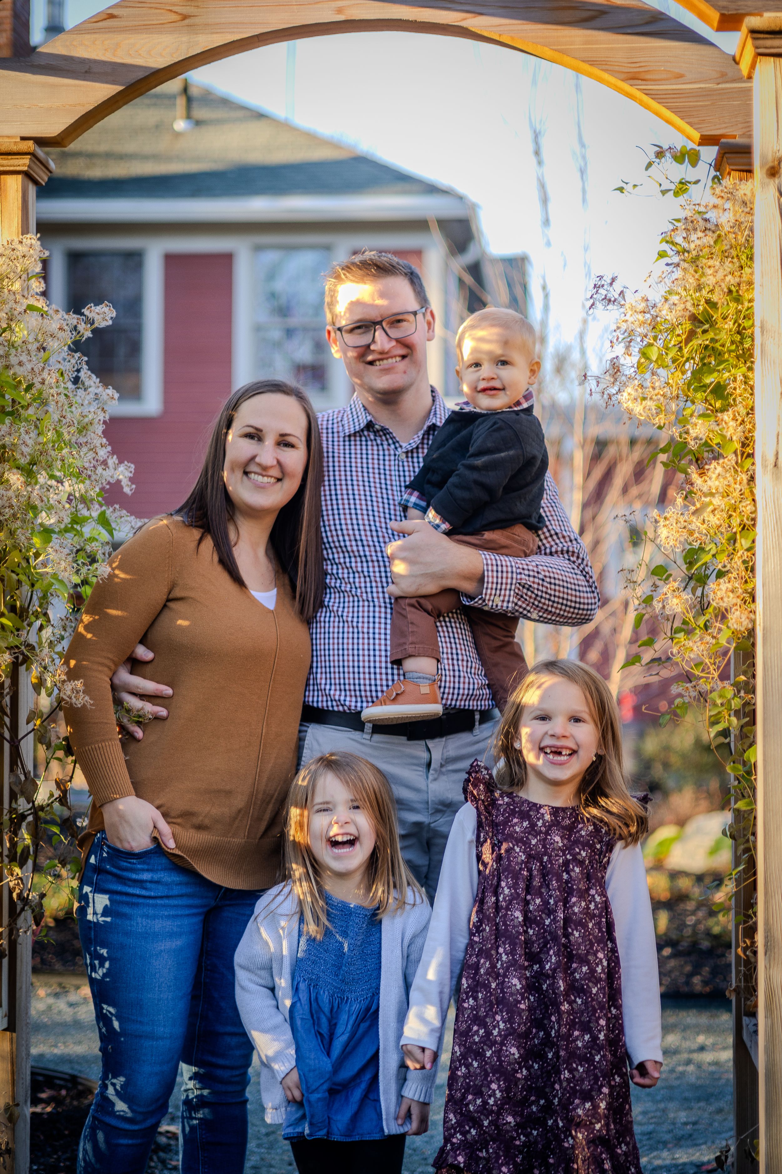 A happy family poses together under an outdoor arch surrounded by autumn foliage and flowers.