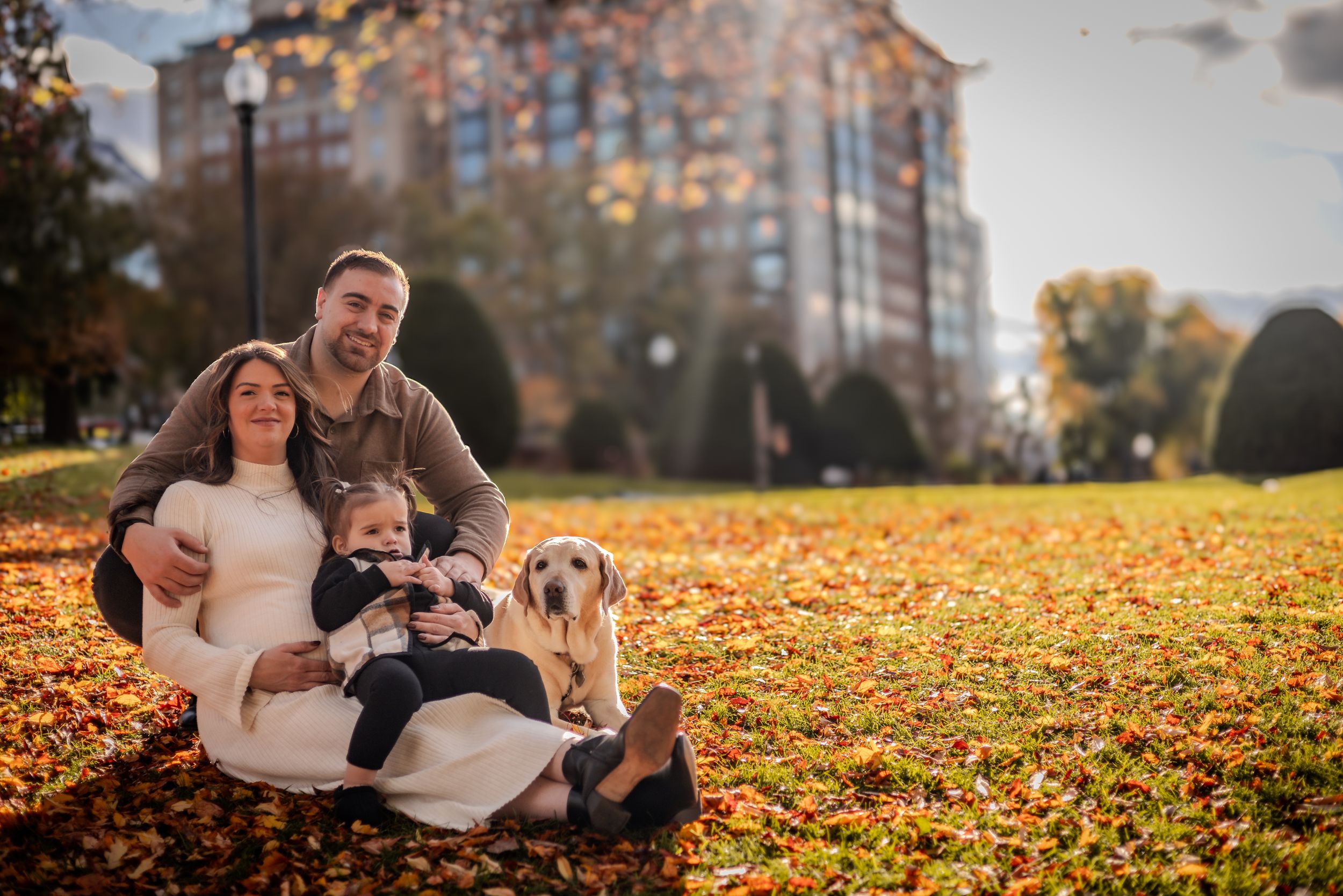 A family sits with their dog on fallen autumn leaves in a park with buildings in the background at sunset.