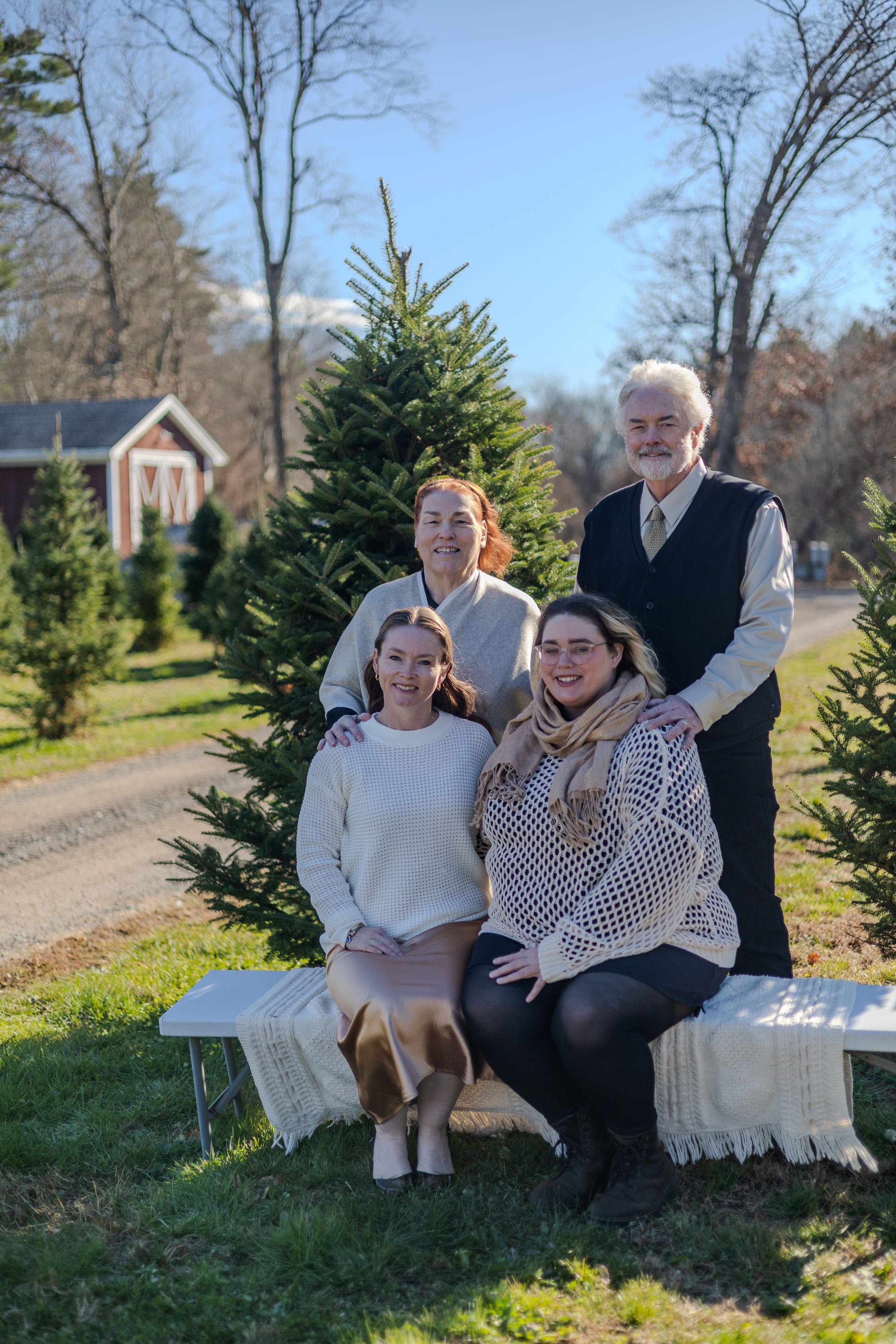 A family poses together on outdoor benches at a Christmas tree farm on a sunny winter day.