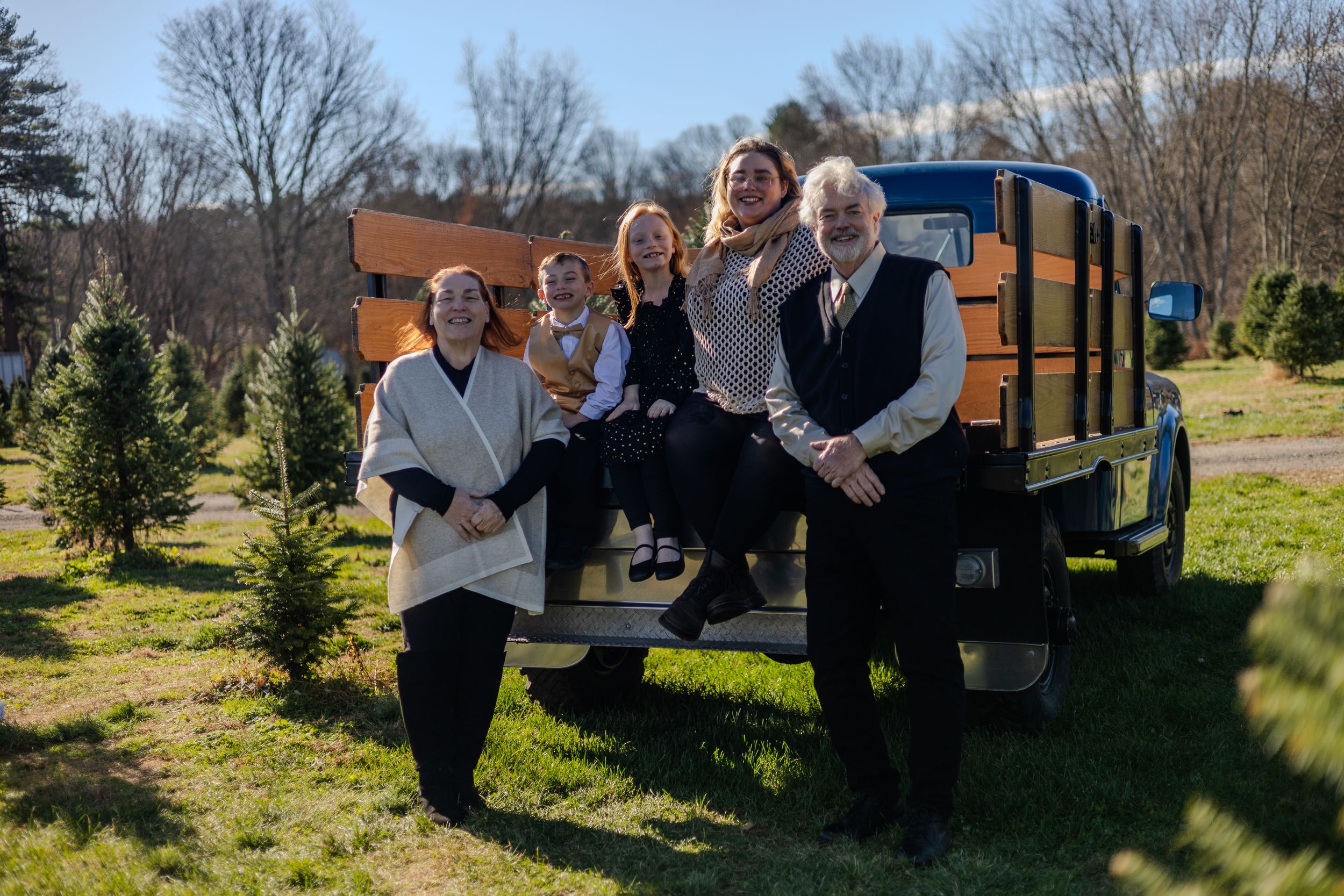 A group of people pose together by a vintage school bus parked among evergreen trees on a sunny day.