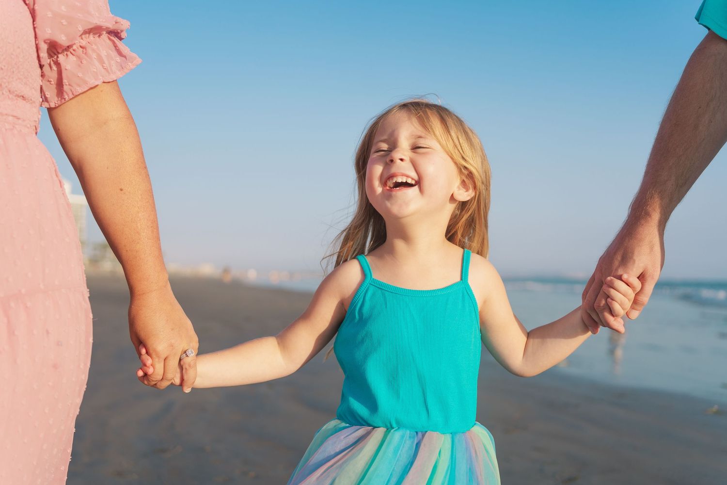 Child wearing a turquoise dress laughs joyfully while holding hands with adults on a sunny beach.
