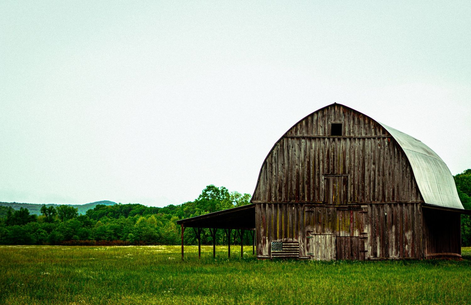 Weathered wooden barn with curved roof standing in green pasture under cloudy sky.