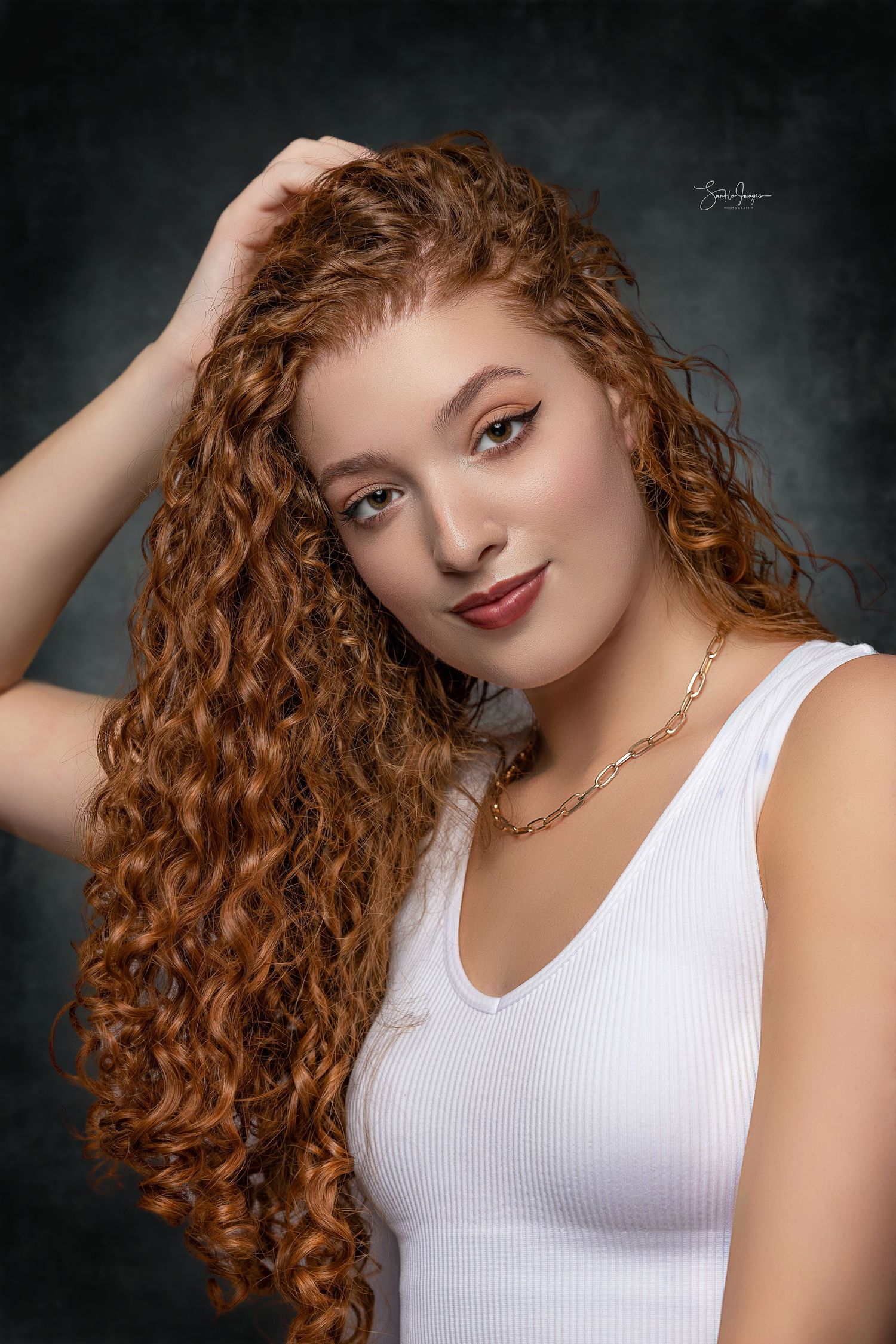 Studio portrait with dramatic lighting shows long curly red hair and white tank top against dark background.