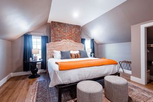 Stylish attic bedroom with exposed brick wall, white bedding, and orange accents on hardwood floors.