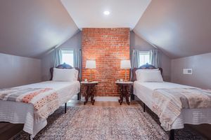 Twin bedroom with exposed brick wall, vaulted ceiling, and matching white beds on a patterned area rug.