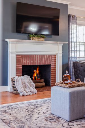 Elegant living room with brick fireplace, mounted TV, and gray ottoman beside a glowing fire.