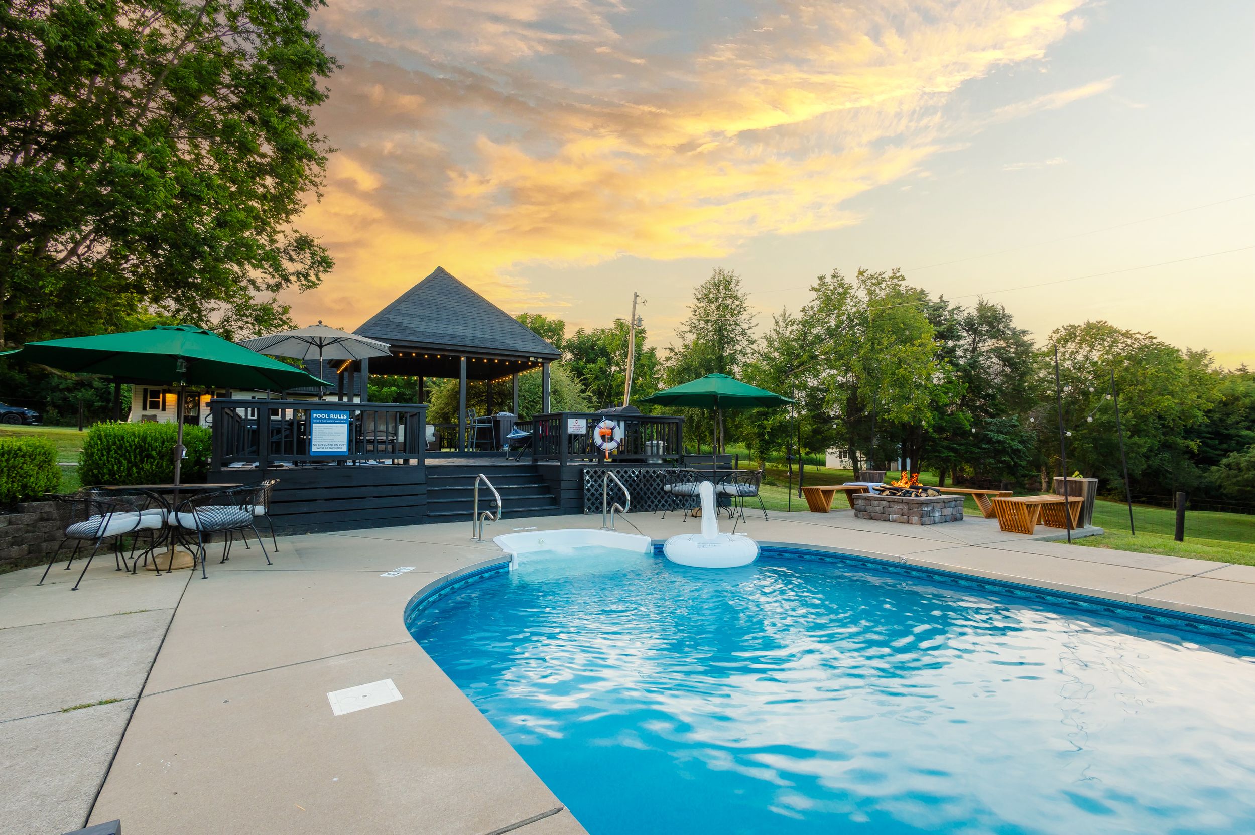 A panoramic view of a swimming pool with lounge chairs against a golden sunset sky and surrounding trees.