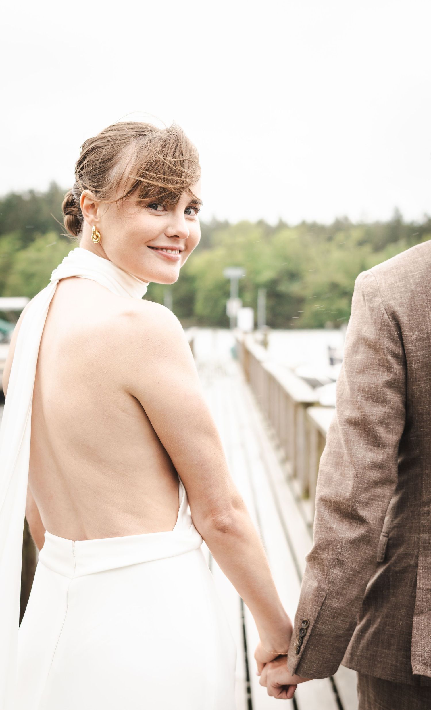 Bride in backless white halter gown smiles over shoulder while holding hands with groom in brown suit on waterfront dock.