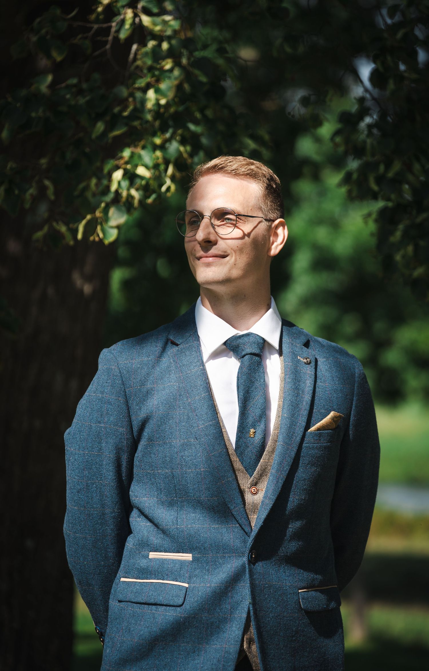 Professional wedding portrait showing groom in blue suit with brown tie and pocket square against natural outdoor setting.