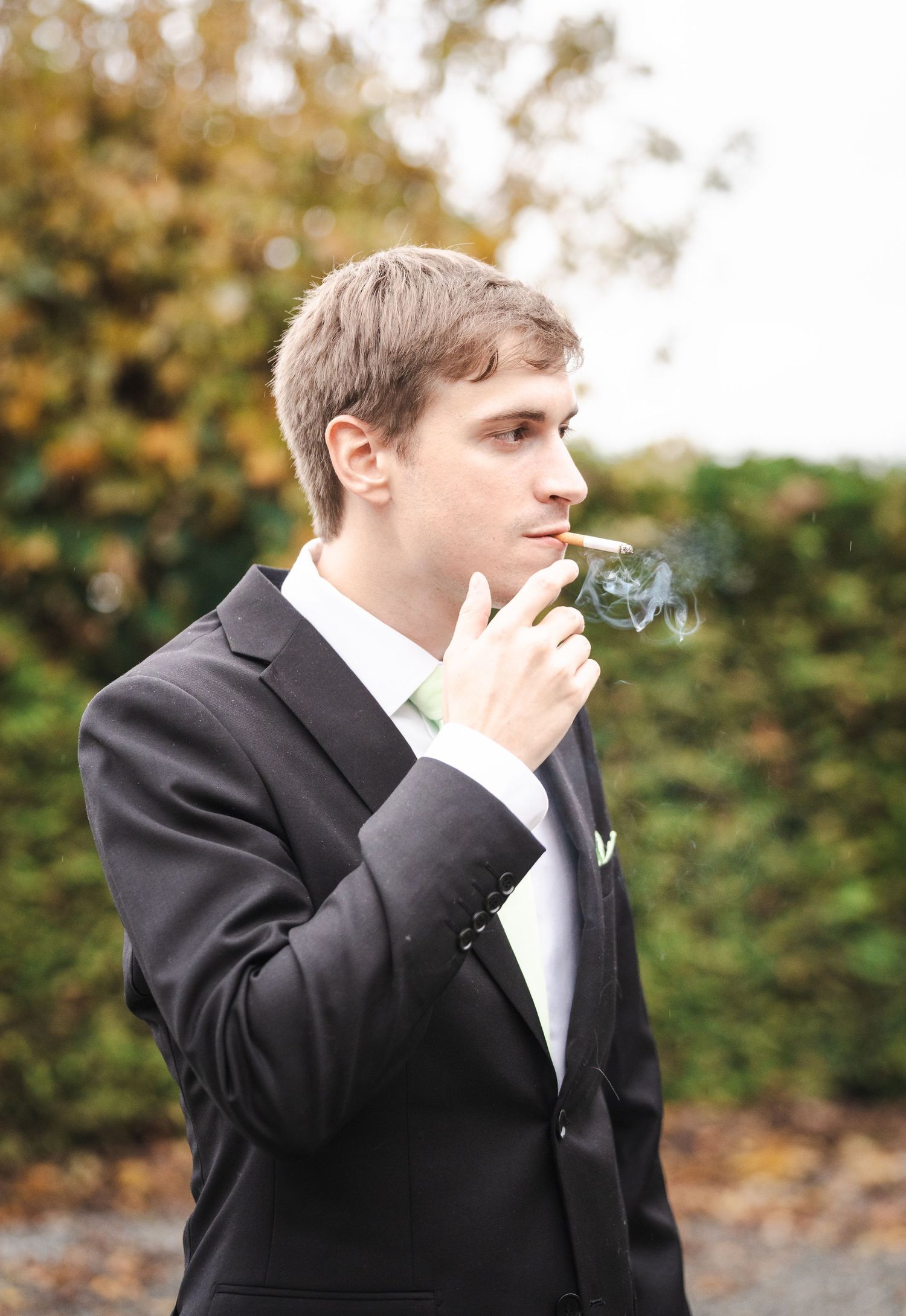 Person in formal black suit and white tie smoking a cigarette outdoors with autumn foliage in background.