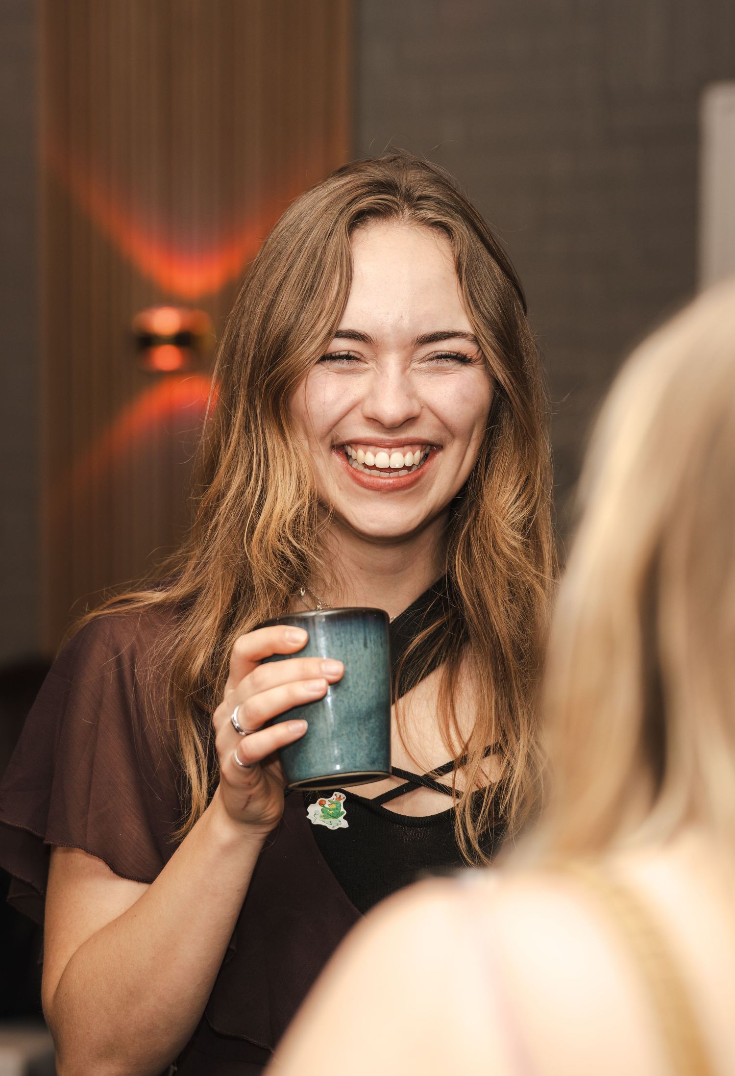 A person with long brown hair laughs joyfully while holding a turquoise coffee mug in a dimly lit setting.