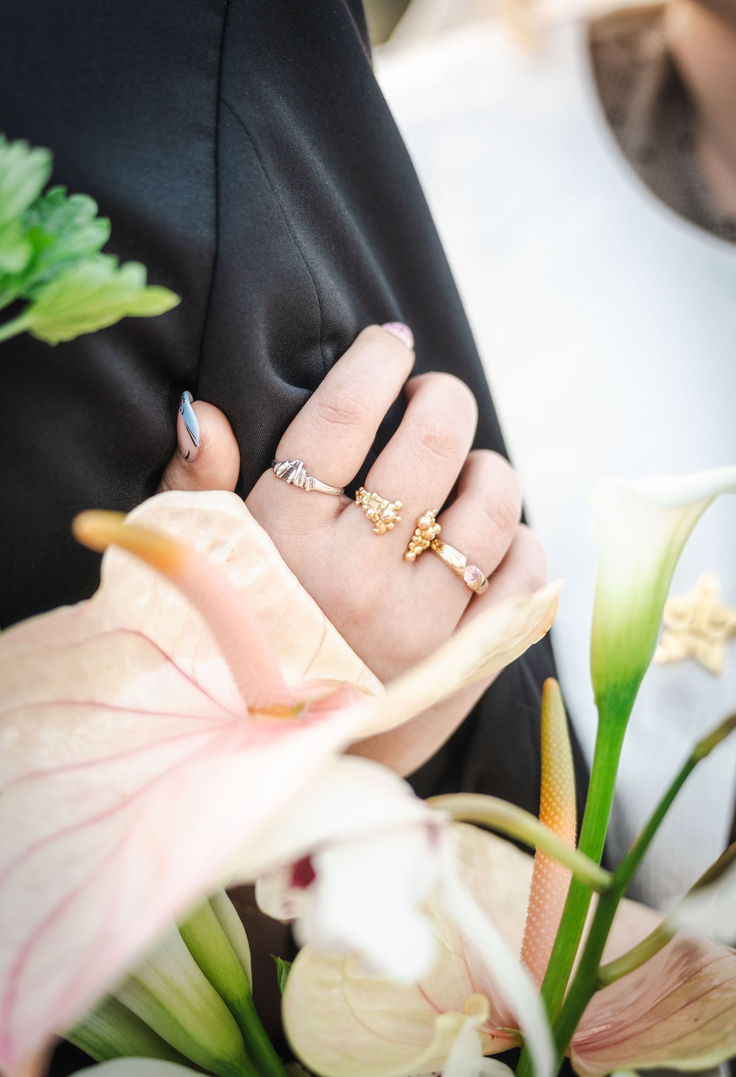 A hand wearing delicate gold rings is shown against white lilies and greenery in soft natural lighting.