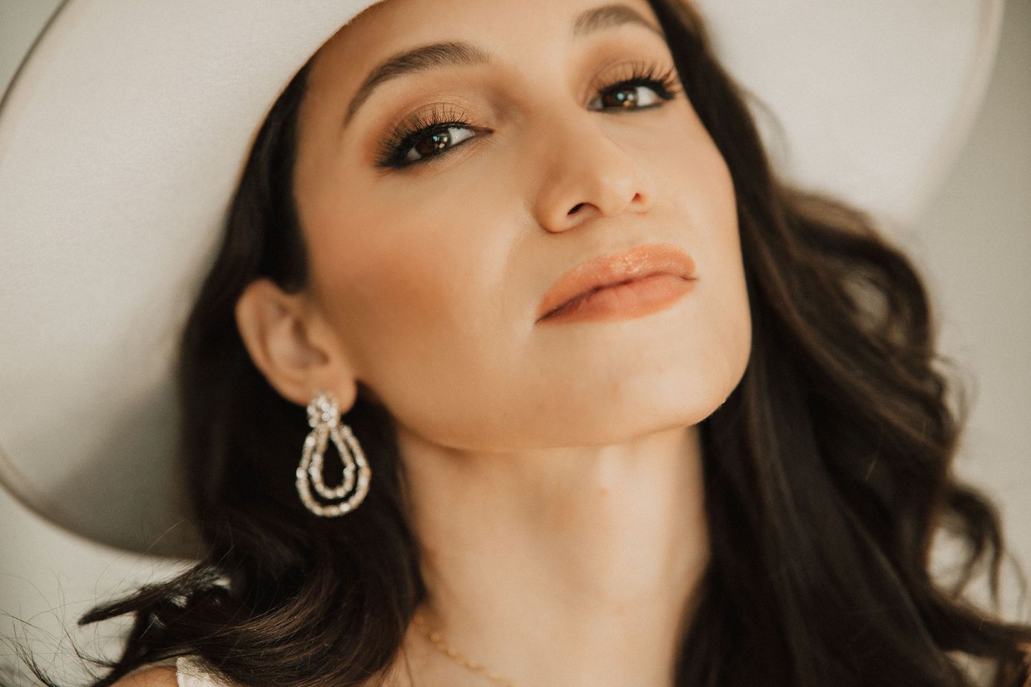A close-up portrait of someone in a white hat with teardrop earrings and dark wavy hair against a light background.
