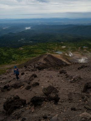 Rocky mountain summit trail with distant mountain ranges and valleys visible through cloudy skies.