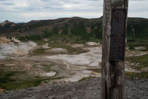 Wooden trail markers stand against a mountainous backdrop with geothermal features.