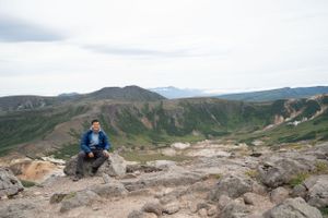 Rocky mountain trail overlooks a vast geothermal landscape with distant peaks.