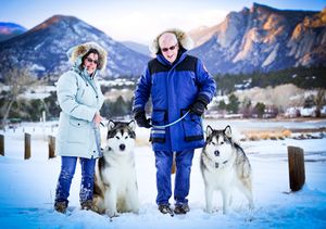 Couple standing with two Alaskan Malamute dogs in snowy mountain landscape.