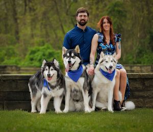 A couple poses with three large Alaskan Malamutes wearing blue bandanas in a park setting.