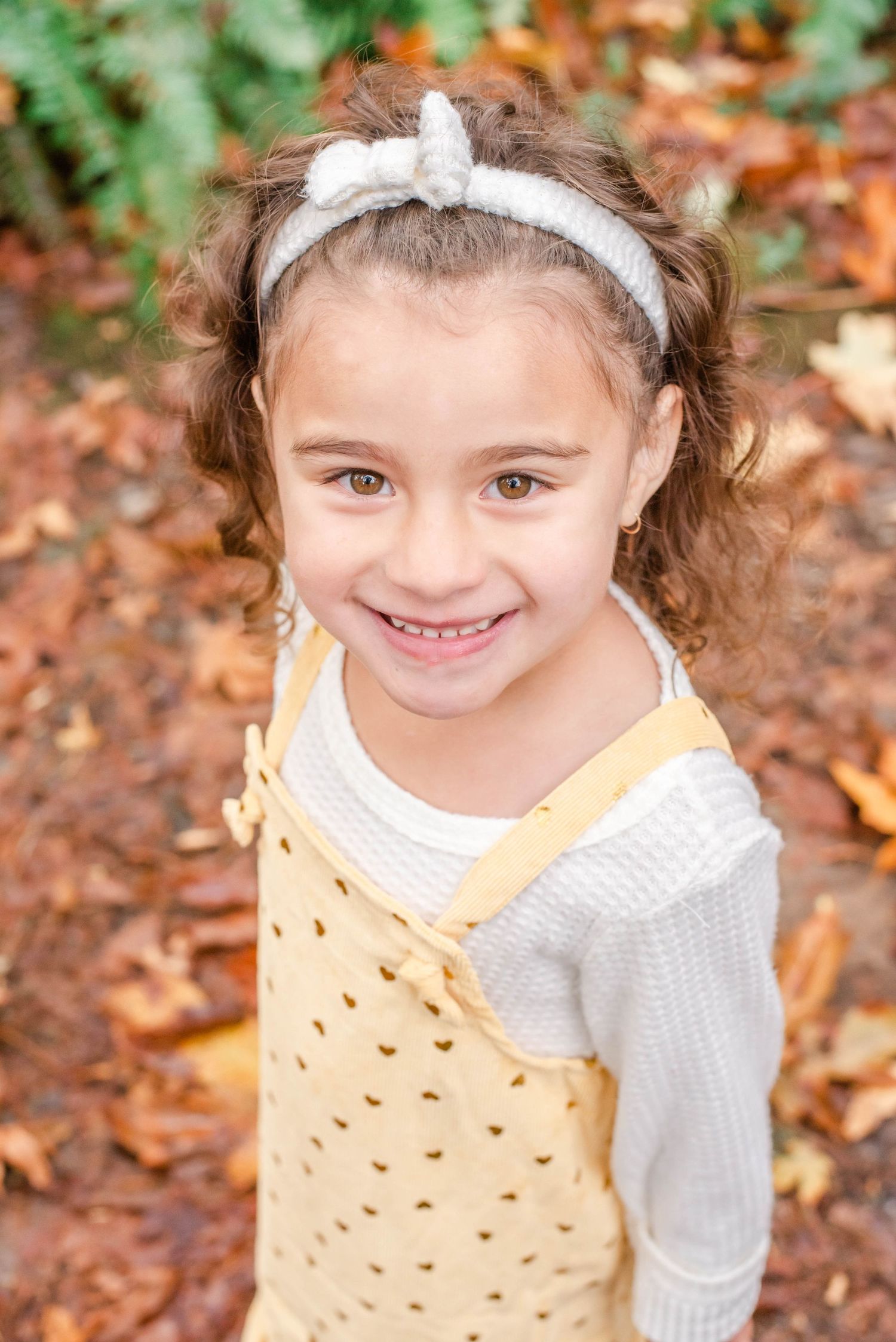 A joyful autumn portrait wearing a yellow jumper dress and white bow headband amidst fallen leaves.