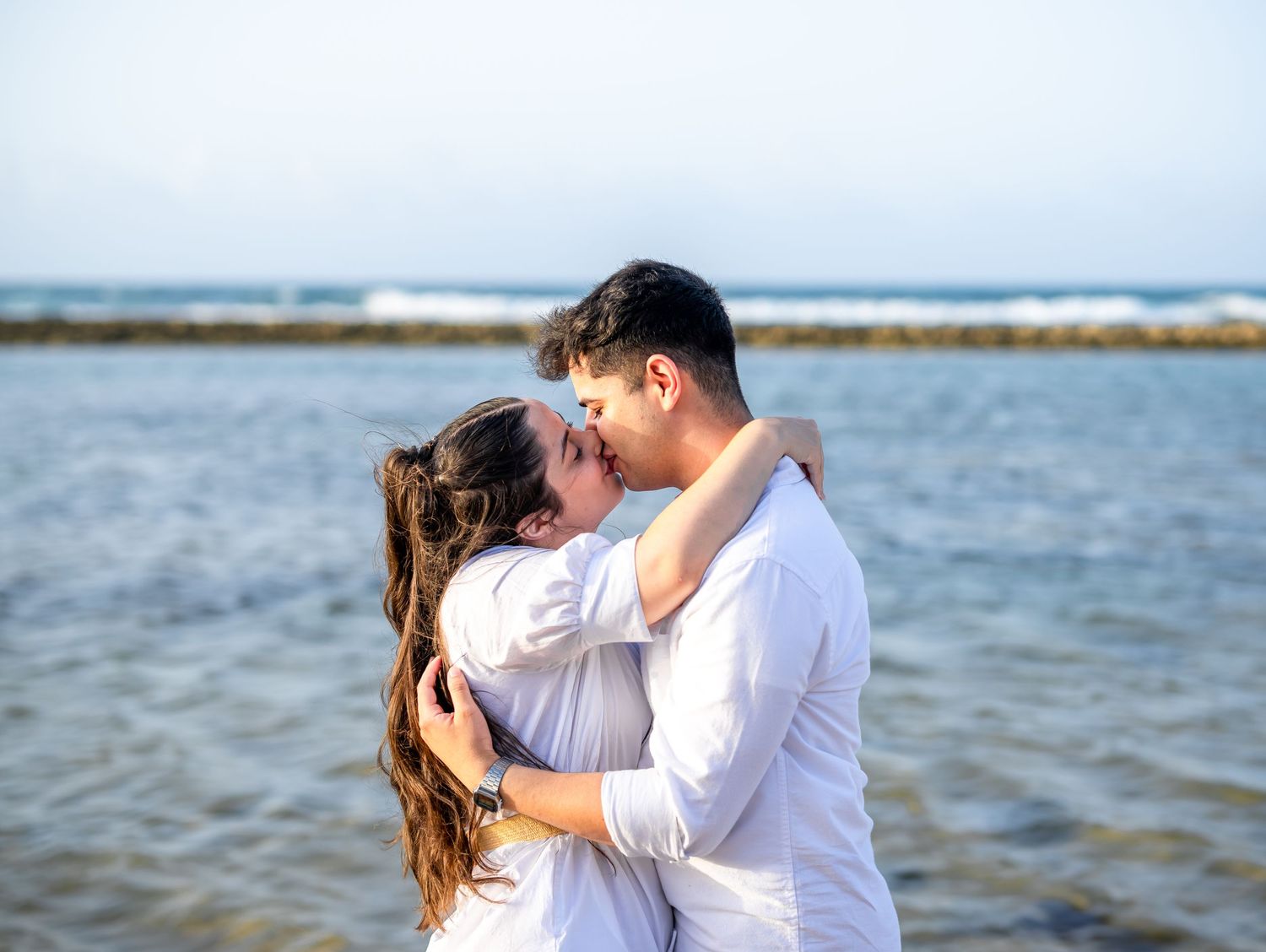 Romantic couple sharing an intimate kiss on a sunny beach with ocean waves in the background.