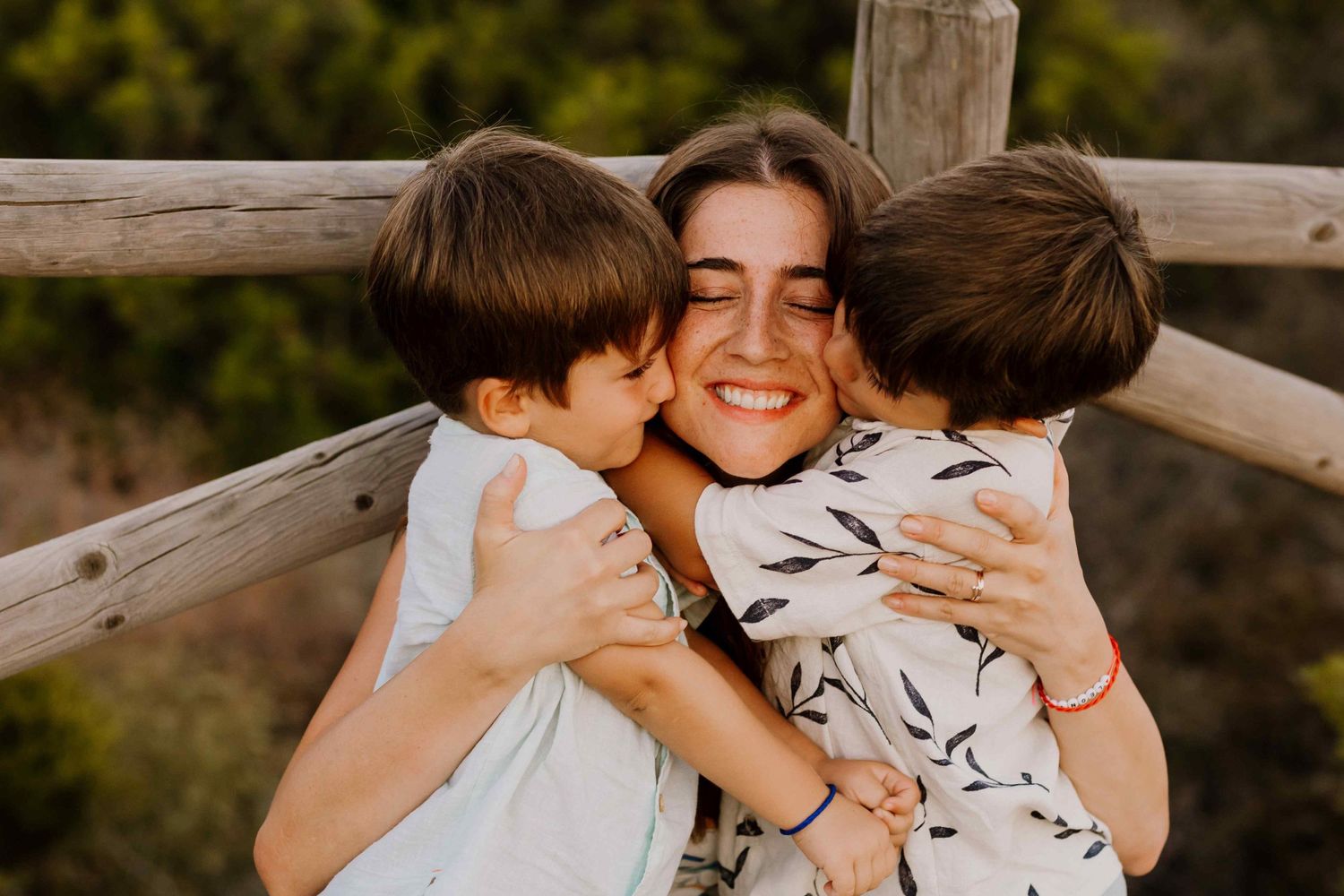 A joyful mother with freckles and a big smile, receiving kisses on both cheeks from her two young sons while hugging them tightly outdoors.