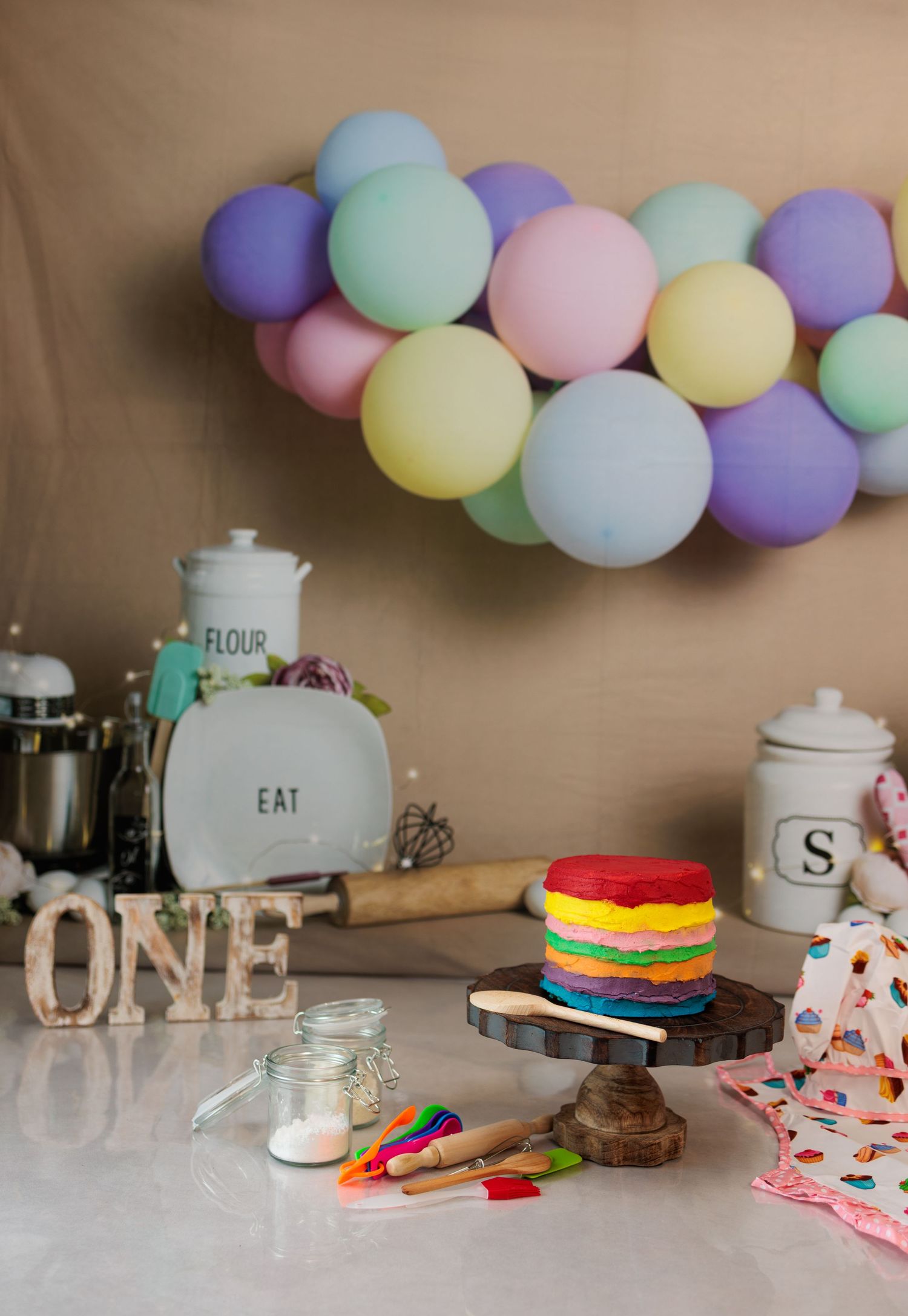 Colorful rainbow cake on a stand with pastel balloons and ONE sign for a first birthday.