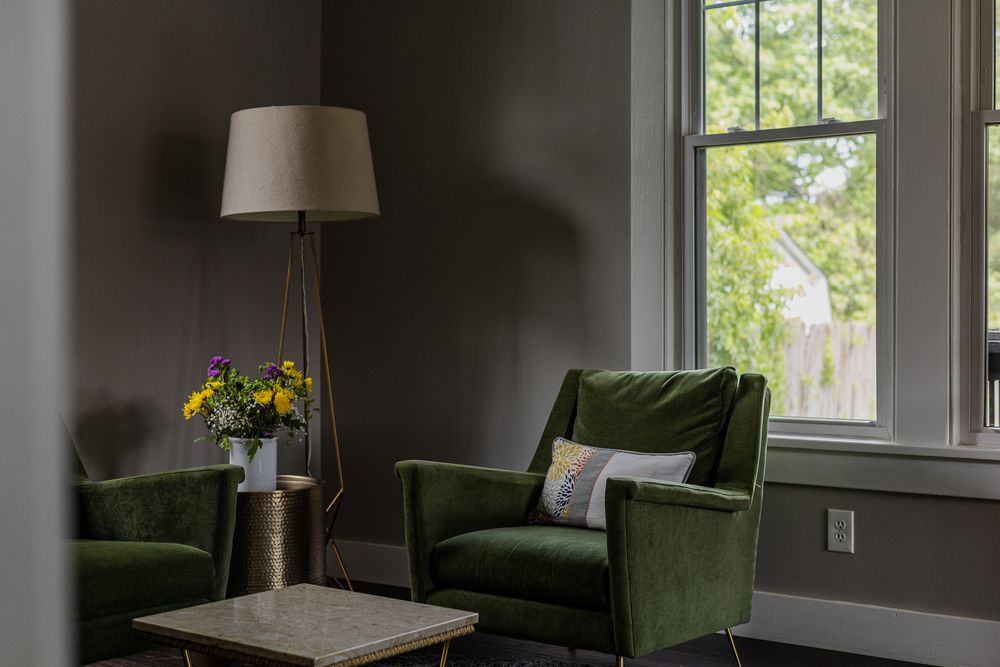 Cozy reading nook with green velvet armchairs, brass floor lamp, marble coffee table, and yellow flowers by a bright window.
